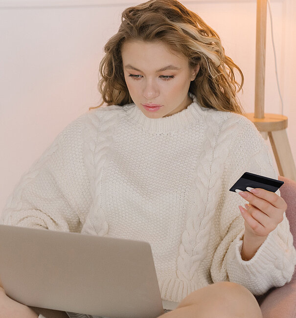 Frau mit blonden langen Haaren sitzt im weißen Wollpulli vor einem Laptop und hält in der Hand eine Kreditkarte