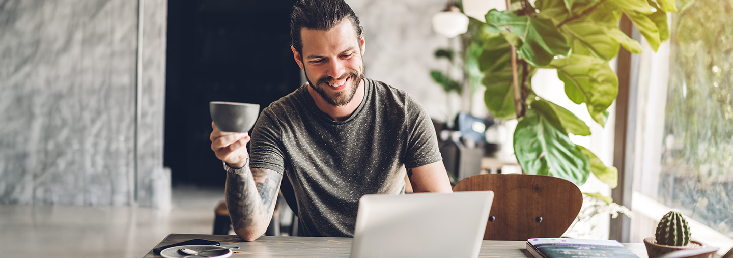 Junger, lachender Mann mit Kaffeetasse vor Laptop